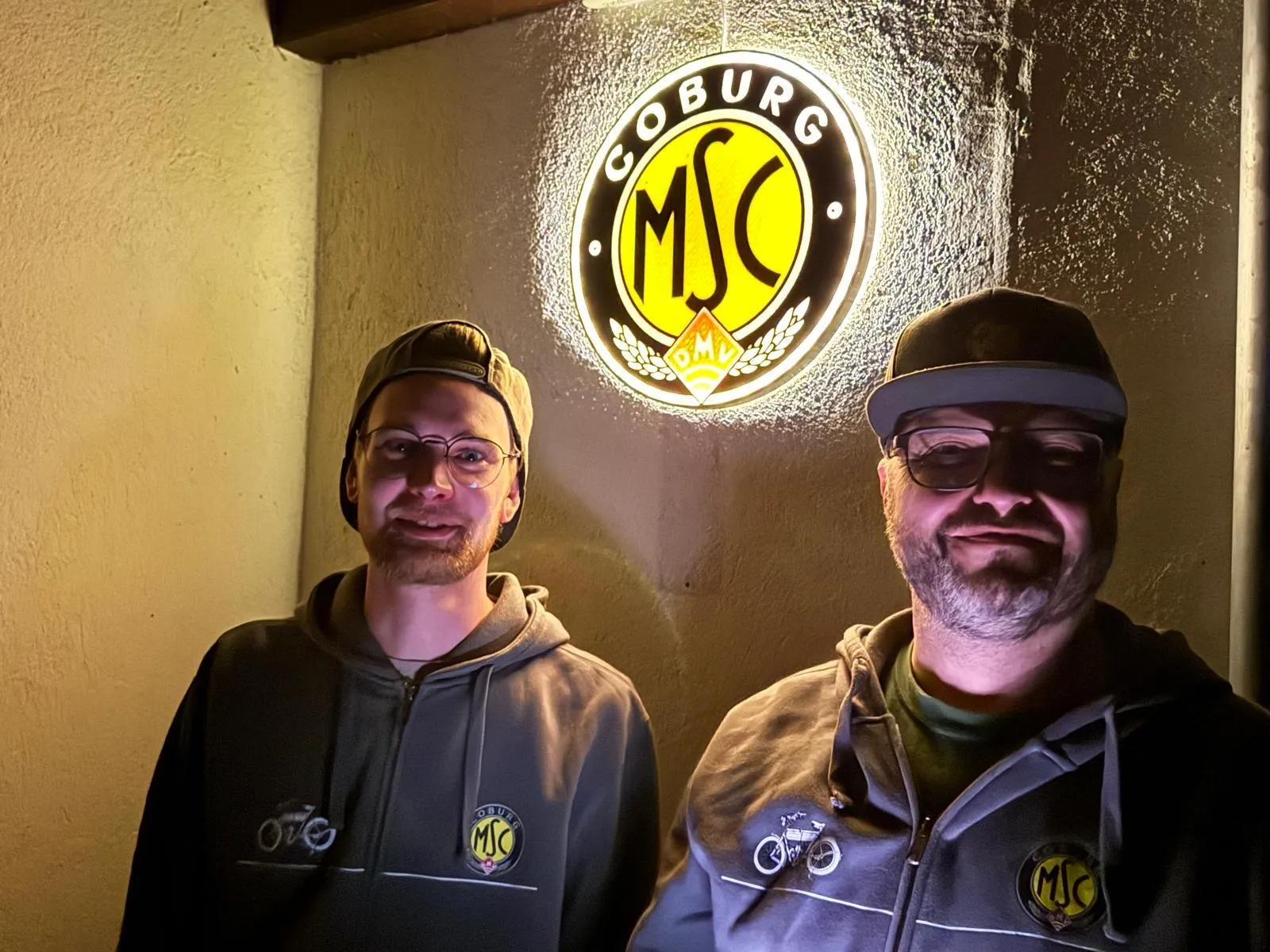 Zwei MSC-Mitglieder unter dem beleuchteten Vereinswappen